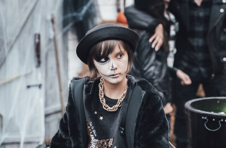 Beautiful scary little girl celebrating halloween. Terrifying black, white half-face makeup and witch costume, stylish image. Horror, fun at children's party in barn on street. Hat, fur coat, chain.の写真素材