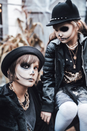Beautiful scary little girl celebrating halloween. Terrifying black, white half-face makeup, witch costume, stylish image. Horror, fun at children's party in barn on street. Hat, jacket.の写真素材