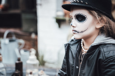 Beautiful scary little girl celebrating halloween. Terrifying black, white half-face makeup, witch costume, stylish image. Horror, fun at children's party in barn on street. Hat, jacket.の写真素材