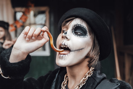 Beautiful scary little girl celebrating halloween, chewing gummy worm. Terrifying face makeup and...