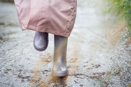 Little girl in pink waterproof raincoat, purple rubber boots funny jumps through puddles on street...
