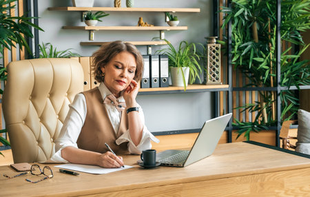 Confident stylish mature middle aged elderly woman in office.Workplace with laptop,table,coffee,glasses. General manager,ceo.Old senior businesswoman,60s lady in suit. Bank business career.の写真素材