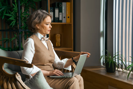 Confident stylish mature middle aged elderly woman in office.Workplace with laptop,using talking by mobile phone.General manager,ceo.Old senior businesswoman,60s lady in suit.Bank entrepreneurial career.の写真素材