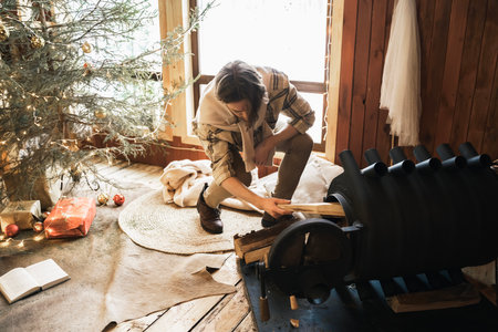 Young stylish man is stoking stove.Celebrating new year eve in cozy wooden country house in winter forest.Gifts,family holiday.Romantic weekend for love couple.Christmas tree with gold red xmas balls.の写真素材