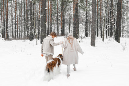 Love romantic young couple girl, guy in snowy cold winter forest walking with pet, dog of hunting breed russian borzoi. Sighthound, wolfhound owner. Having fun, laughing. Stylish fur coat, woolen hat.の写真素材