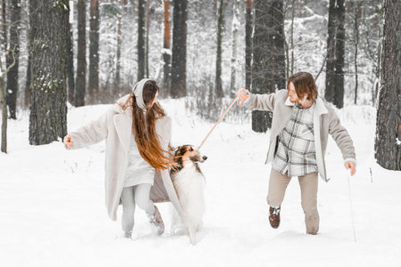 Love romantic young couple girl, guy in snowy cold winter forest walking with pet, dog of hunting breed russian borzoi. Sighthound, wolfhound owner. Having fun, laughing. Stylish fur coat, woolen hat.の写真素材