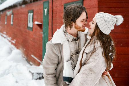 Young couple girl guy walking near barn with skates. Snowy cold winter in the countryside. Skating, kissing, having fun, laughing in stylish clothes, fur coat, hat. Romantic love story, village date, weekend.の写真素材
