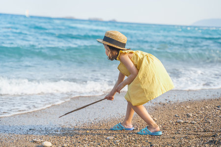 Little girl walking on beach, sea ocean shore in romantic yellow dress, straw hat. Playing in sand, blue waves. Family vacation travel leisure in hot summer coast. Sunny day relax in hotel resort.の写真素材