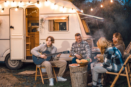 Family trailer travel.Children,brother sister,mom dad playing guitar,singing song at fire.Evening picnic in nature.Holiday barbecue BBQ food.Vacation weekend dinner.Road lunch.Camper,house on wheels.の写真素材