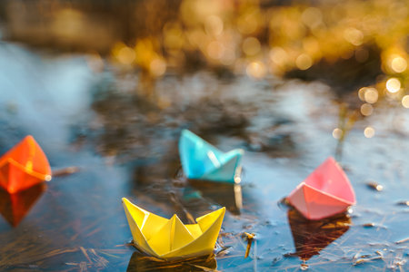 Multicolored paper boats. Colorful pink yellow blue orange ships in big spring snow puddle on winter street. Warm wet rainy weather, old grass. hello spring autumn. Children play, have fun outdoors.の写真素材