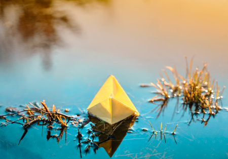 Yellow floating paper boat. Colorful sailing ship in big blue spring puddle, river water on winter street. Warm wet rainy weather, old grass. hello spring autumn. Children play, have fun outdoors.の写真素材