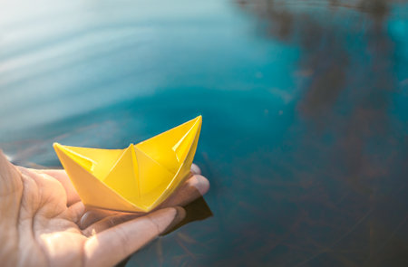 Yellow lonely floating paper boat in hand. Colorful sailing ship in big blue spring puddle, river water.Warm wet rainy weather.Hello spring,autumn.Children play,have street fun outdoors.Way to future.の写真素材