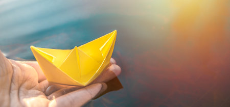 Yellow lonely floating paper boat in hand. Colorful sailing ship in big blue spring puddle, river water.Warm wet rainy weather.Hello spring,autumn.Children play,have street fun outdoors.Way to future.の写真素材