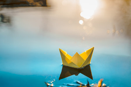 Yellow floating paper boat. Colorful sailing ship in big blue spring puddle, river water on winter street. Warm wet rainy weather, old grass. hello spring autumn. Children play, have fun outdoors.の写真素材