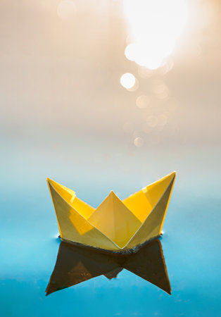 Yellow floating paper boat. Colorful sailing ship in big blue spring puddle, river water on winter street. Warm wet rainy weather, old grass. hello spring autumn. Children play, have fun outdoors.の写真素材