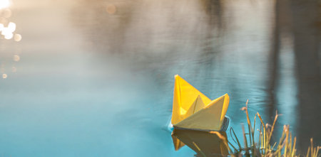Yellow floating paper boat. Colorful sailing ship in big blue spring puddle, river water on winter street. Warm wet rainy weather, old grass. hello spring autumn. Children play, have fun outdoors.の写真素材
