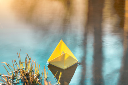Yellow floating paper boat. Colorful sailing ship in big blue spring puddle, river water on winter street. Warm wet rainy weather, old grass. hello spring autumn. Children play, have fun outdoors.の写真素材