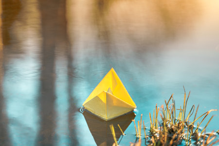 Yellow floating paper boat. Colorful sailing ship in big blue spring puddle, river water on winter street. Warm wet rainy weather, old grass. hello spring autumn. Children play, have fun outdoors.の写真素材