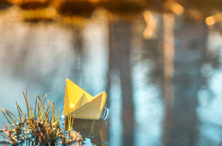 Yellow floating paper boat. Colorful sailing ship in big blue spring puddle, river water on winter street. Warm wet rainy weather, old grass. hello spring autumn. Children play, have fun outdoors.の写真素材