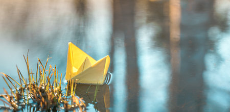 Yellow floating paper boat. Colorful sailing ship in big blue spring puddle, river water on winter street. Warm wet rainy weather, old grass. hello spring autumn. Children play, have fun outdoors.の写真素材