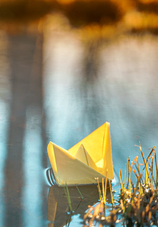 Yellow floating paper boat. Colorful sailing ship in big blue spring puddle, river water on winter street. Warm wet rainy weather, old grass. hello spring autumn. Children play, have fun outdoors.の写真素材