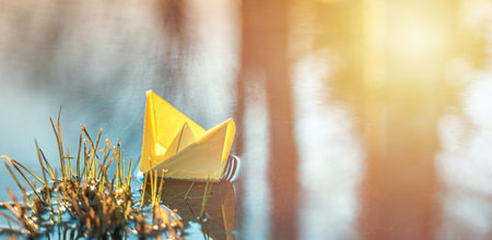 Yellow floating paper boat. Colorful sailing ship in big blue spring puddle, river water on winter street. Warm wet rainy weather, old grass. hello spring autumn. Children play, have fun outdoors.の写真素材
