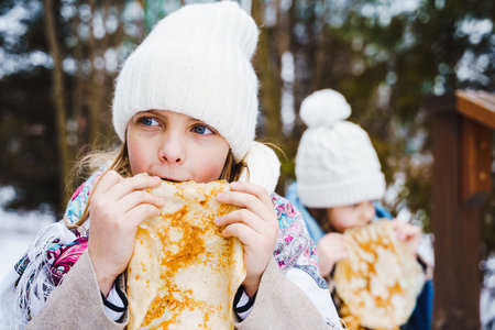 Russian national festival Maslenitsa, shrovetide. Little cute beautiful girls in headscarf eat big tasty pancakes, have fun on winter pancake holiday week. Traditional folf slavic street carnival.の写真素材