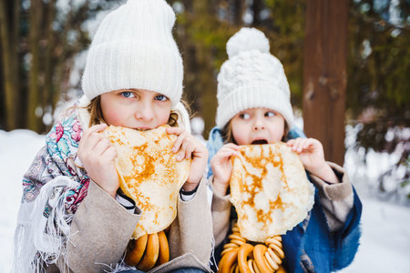 Russian national festival Maslenitsa, shrovetide. Little cute beautiful girls in headscarf eat big tasty pancakes, have fun on winter pancake holiday week. Traditional folf slavic street carnival.の写真素材
