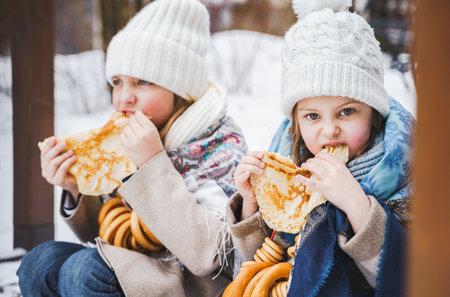 Russian national festival Maslenitsa, shrovetide. Little cute beautiful girls in headscarf eat big tasty pancakes, have fun on winter pancake holiday week. Traditional folf slavic street carnival.の写真素材