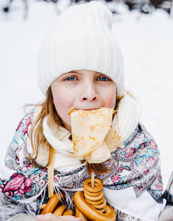 Russian national festival Maslenitsa, shrovetide. Little cute beautiful girls in headscarf eat big tasty pancakes, have fun on winter pancake holiday week. Traditional folf slavic street carnival.の写真素材