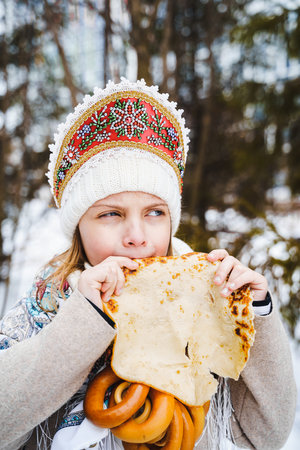 Russian national festival Maslenitsa, shrovetide. Little cute beautiful girls in headscarf eat big tasty pancakes, have fun on winter pancake holiday week. Traditional folf slavic street carnival.の写真素材