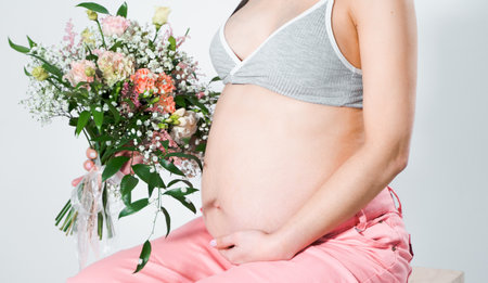 Pregnant woman with bouquet of flowers. Girl in gray top, pink pants holds hands on naked belly. Photoshoot of happy pregnancy, maternity, preparation. baby expectation. Beautiful tender young mother.の写真素材