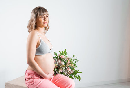 Pregnant woman with bouquet of flowers. Girl in gray top, pink pants holds hands on naked belly. Photoshoot of happy pregnancy, maternity, preparation. baby expectation. Beautiful tender young mother.の写真素材