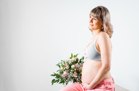 Pregnant woman with bouquet of flowers. Girl in gray top, pink pants holds hands on naked belly. Photoshoot of happy pregnancy, maternity, preparation. baby expectation. Beautiful tender young mother.の写真素材