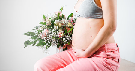 Pregnant woman with bouquet of flowers. Girl in gray top, pink pants holds hands on naked belly. Photoshoot of happy pregnancy, maternity, preparation. baby expectation. Beautiful tender young mother.の写真素材