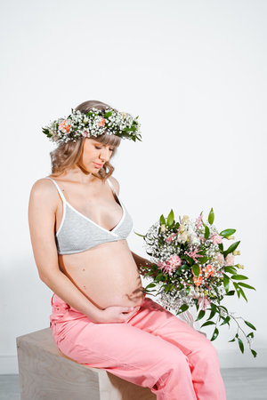 Pregnant woman in wreath with bouquet of flowers. Girl in gray top, pink pants. Hands on naked belly. Photoshoot happy pregnancy,maternity,preparation. baby expectation. Beautiful tender young mother.の写真素材