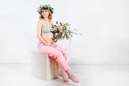 Pregnant woman in wreath with bouquet of flowers. Girl in gray top, pink pants. Hands on naked belly. Photoshoot happy pregnancy,maternity,preparation. baby expectation. Beautiful tender young mother.の写真素材