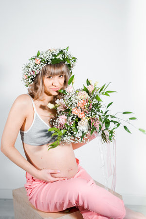 Pregnant woman in wreath with bouquet of flowers. Girl in gray top, pink pants. Hands on naked belly. Photoshoot happy pregnancy,maternity,preparation. baby expectation. Beautiful tender young mother.の写真素材