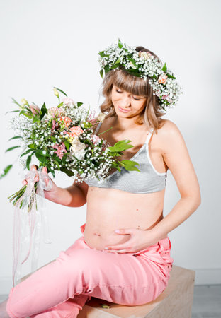 Pregnant woman in wreath with bouquet of flowers. Girl in gray top, pink pants. Hands on naked belly. Photoshoot happy pregnancy,maternity,preparation. baby expectation. Beautiful tender young mother.の写真素材
