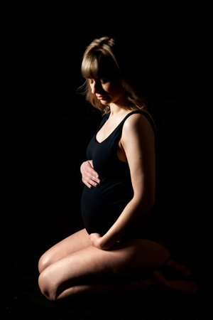 Pregnant woman with big belly in bodysuit, lingerie on black background. Sexy attractive girl photo shoot in studio. Happy pregnancy,maternity,preparation.Baby expectation.Beautiful tender young mother.の写真素材