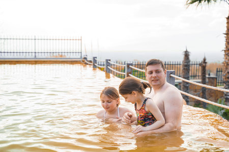 Family, dad and children bathe, swim in outdoor dirty hot springs. Thermal water bath pool and healthy natural, clay and yellow green mud. Spa wellness relax family winter vacation in mountain hotel.の写真素材