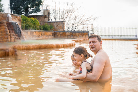 Family, dad and children bathe, swim in outdoor dirty hot springs. Thermal water bath pool and healthy natural, clay and yellow green mud. Spa wellness relax family winter vacation in mountain hotel.の写真素材