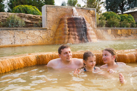Family, dad and children bathe, swim in outdoor dirty hot springs. Thermal water bath pool and healthy natural, clay and yellow green mud. Spa wellness relax family winter vacation in mountain hotel.の写真素材