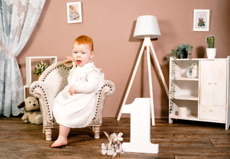 Little redhead baby girl celebrate first birthday anniversary. 1 year family party Professional photoshoot in photo studio. Cute adorable red hair kid in white vintage dress. Children's room at home.の写真素材