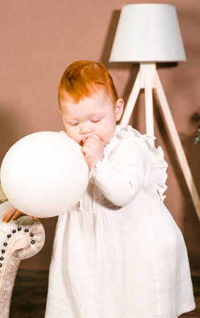 Little redhead baby girl wih balloon celebrates first birthday anniversary. 1 year family party Photoshoot in photo studio. Cute adorable red hair kid in white vintage dress. Children's room at home.の写真素材