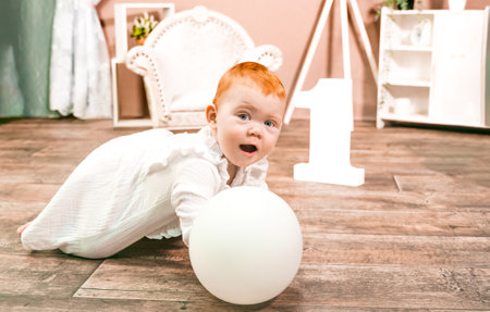 Little redhead baby girl wih balloon celebrates first birthday anniversary. 1 year family party Photoshoot in photo studio. Cute adorable red hair kid in white vintage dress. Children's room at home.の写真素材