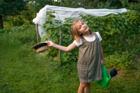 Farmer girl in summer straw hat. Little vegetables gardener farming in garden. Big green watering can water fresh cucumbers seedlings. Harvest help work. Cultivation healthy organic food, countryside.の写真素材