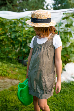 Farmer girl in summer straw hat. Little vegetables gardener farming in garden. Big green watering can water fresh cucumbers seedlings. Harvest help work. Cultivation healthy organic food, countryside.の写真素材