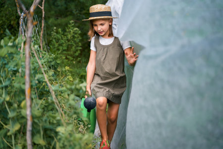 Farmer girl in summer straw hat. Little vegetables gardener farming in garden. Big green watering can water fresh cucumbers seedlings. Harvest help work. Cultivation healthy organic food, countryside.の写真素材