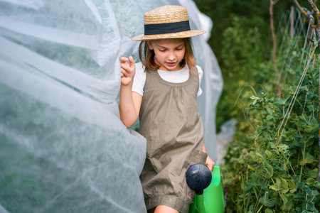 Farmer girl in summer straw hat. Little vegetables gardener farming in garden. Big green watering can water fresh cucumbers seedlings. Harvest help work. Cultivation healthy organic food, countryside.の写真素材